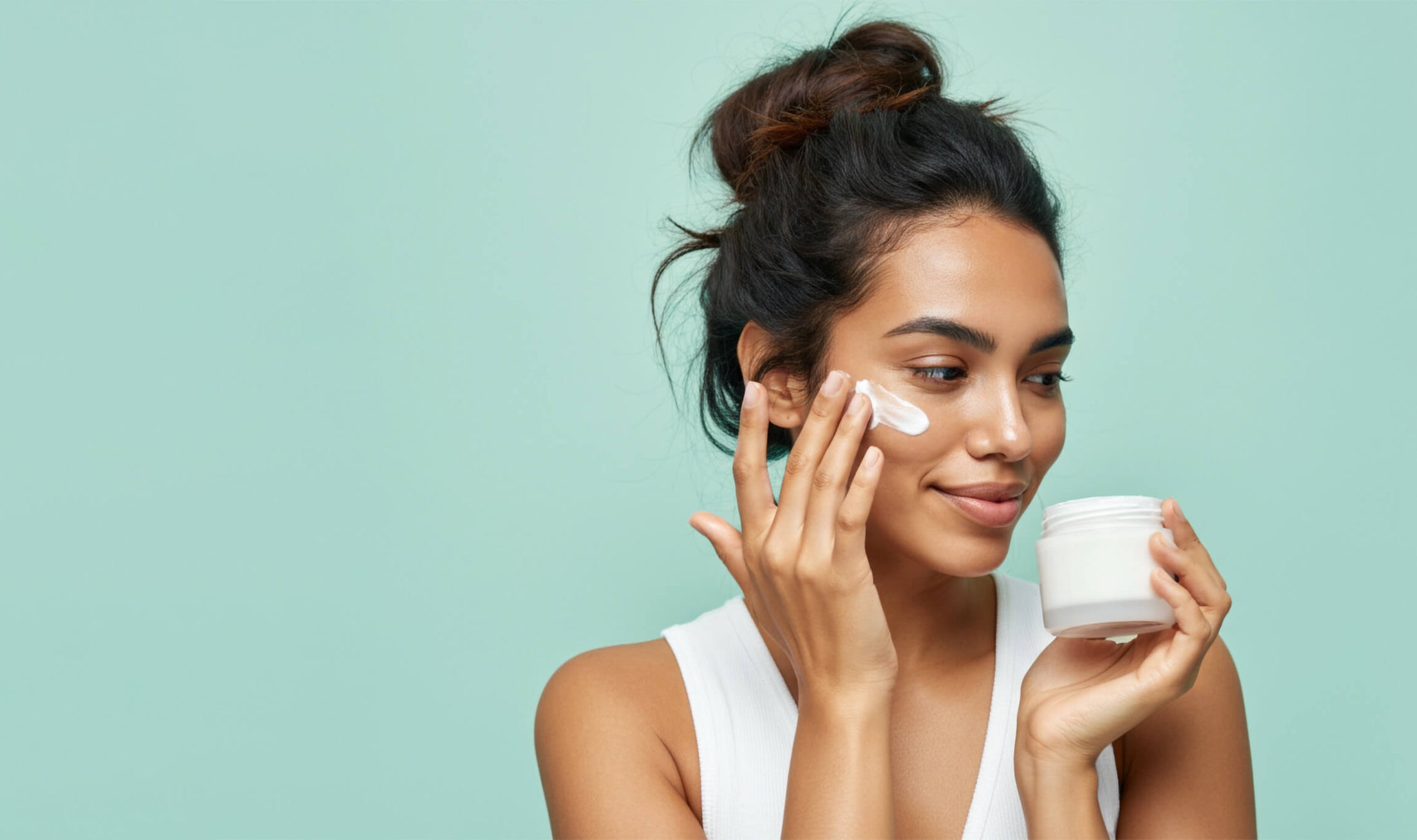 Model against mint green background rubs cream into her cheek, smiling and looking to the side.